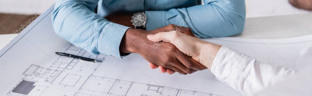 partial view of african american businessman shaking hands with business partner near blueprint, blurred foreground, banner